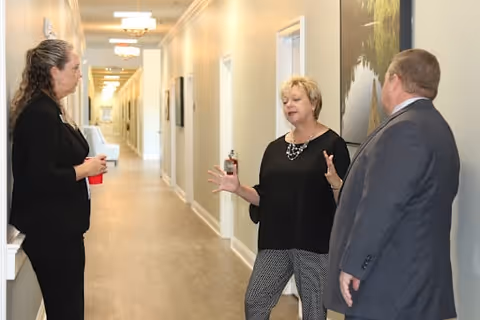 Three adults standing and talking in a well-lit hallway with light-colored walls and wooden flooring. One woman is holding a red cup, another woman is gesturing with her hands, and a man in a suit is listening. The hallway has several doors and framed pictures on the walls.
