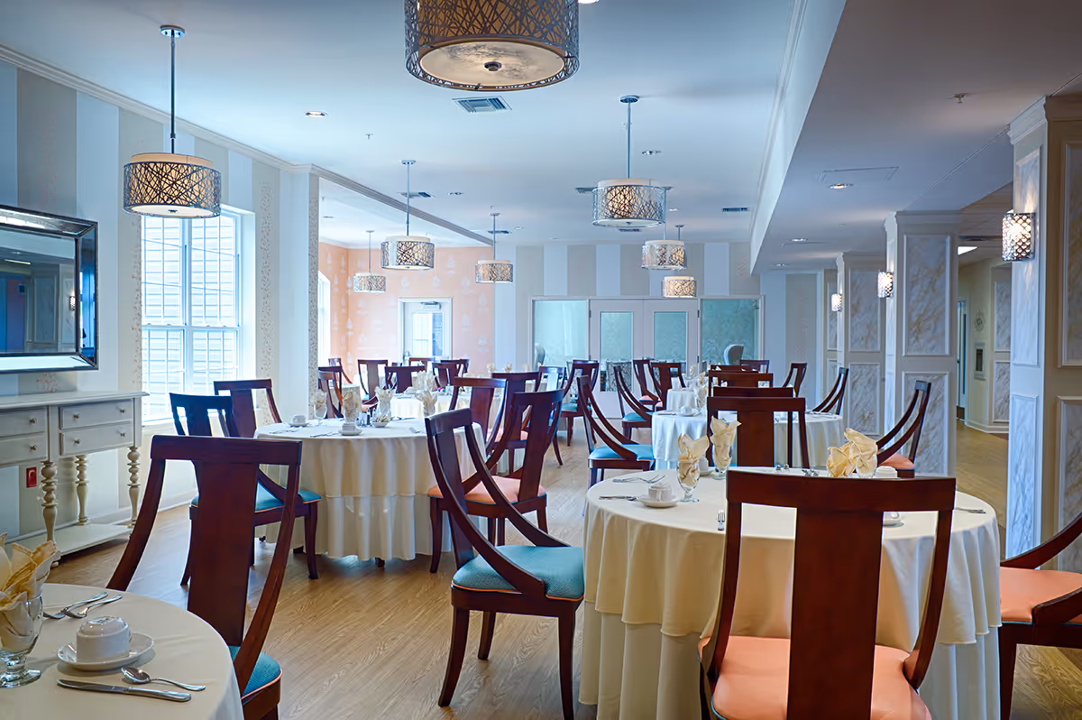 A bright and elegant dining room with round tables covered in white tablecloths, each set with napkins, cups, and silverware. The room features wooden chairs with blue and orange cushions, pendant lights hanging from the ceiling, large windows allowing natural light, and decorative wall panels.