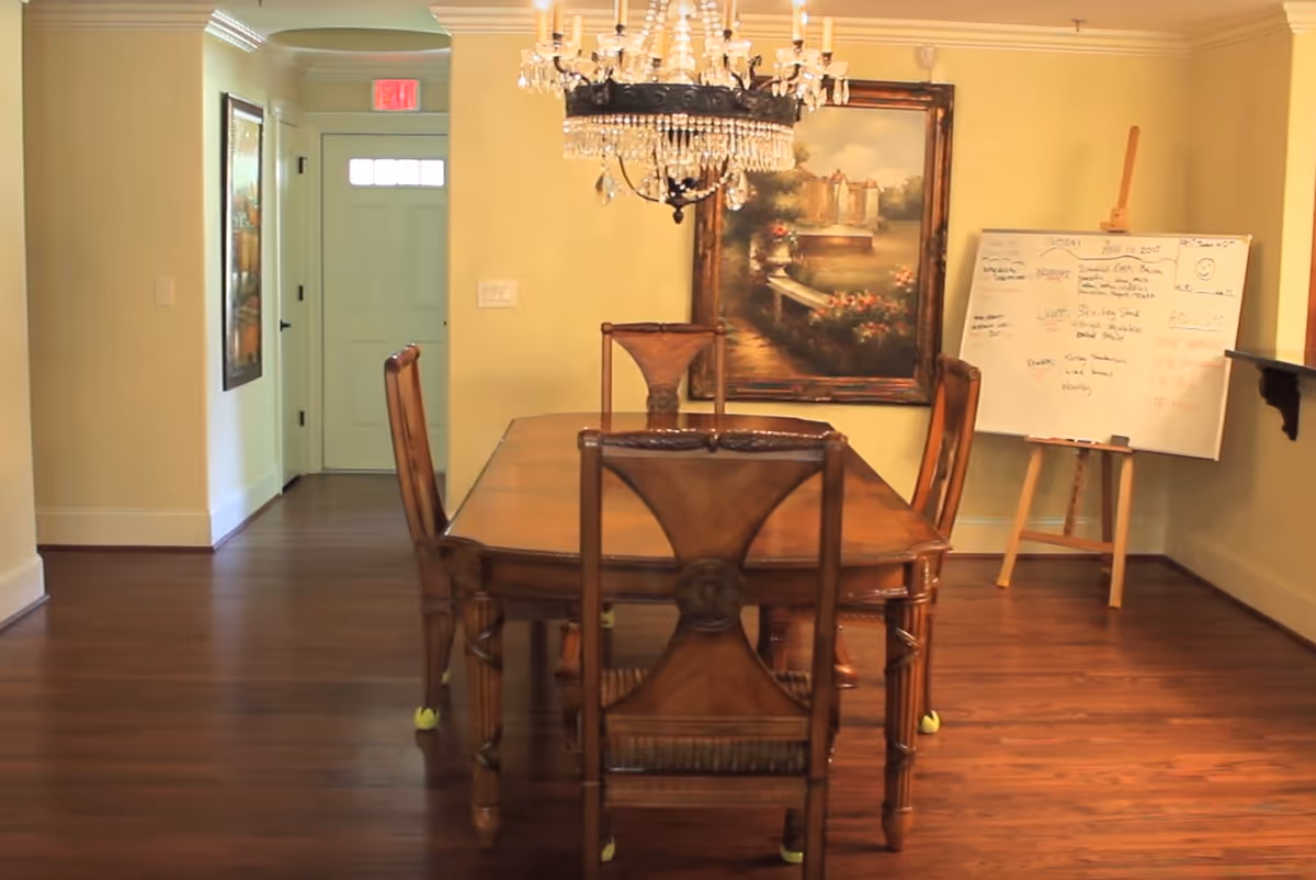 A dining room with a wooden dining table and six chairs, a chandelier hanging above the table, a large framed painting on the wall, and a whiteboard on an easel with notes written on it. The room has wooden flooring and light yellow walls, with a door and hallway visible in the background.