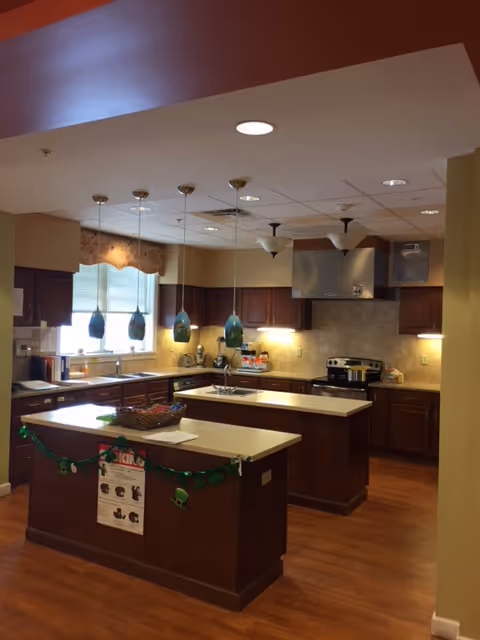 Communal kitchen with two islands, dark wood cabinets, pendant lights, stainless steel range hood, and hardwood floors.