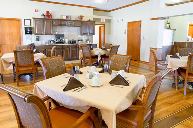Dining area with multiple tables set for meals, wooden chairs, and a kitchenette with cabinets in the background.