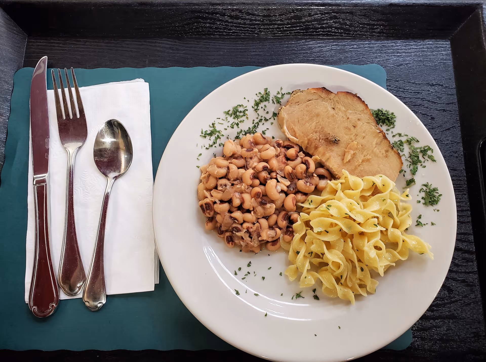 A white plate with a serving of cooked black-eyed peas, a portion of buttered egg noodles, and a slice of roasted meat garnished with chopped herbs. The plate is on a dark wooden tray with a teal placemat underneath. To the left of the plate, there is a folded white napkin with a knife, fork, and spoon placed on top.