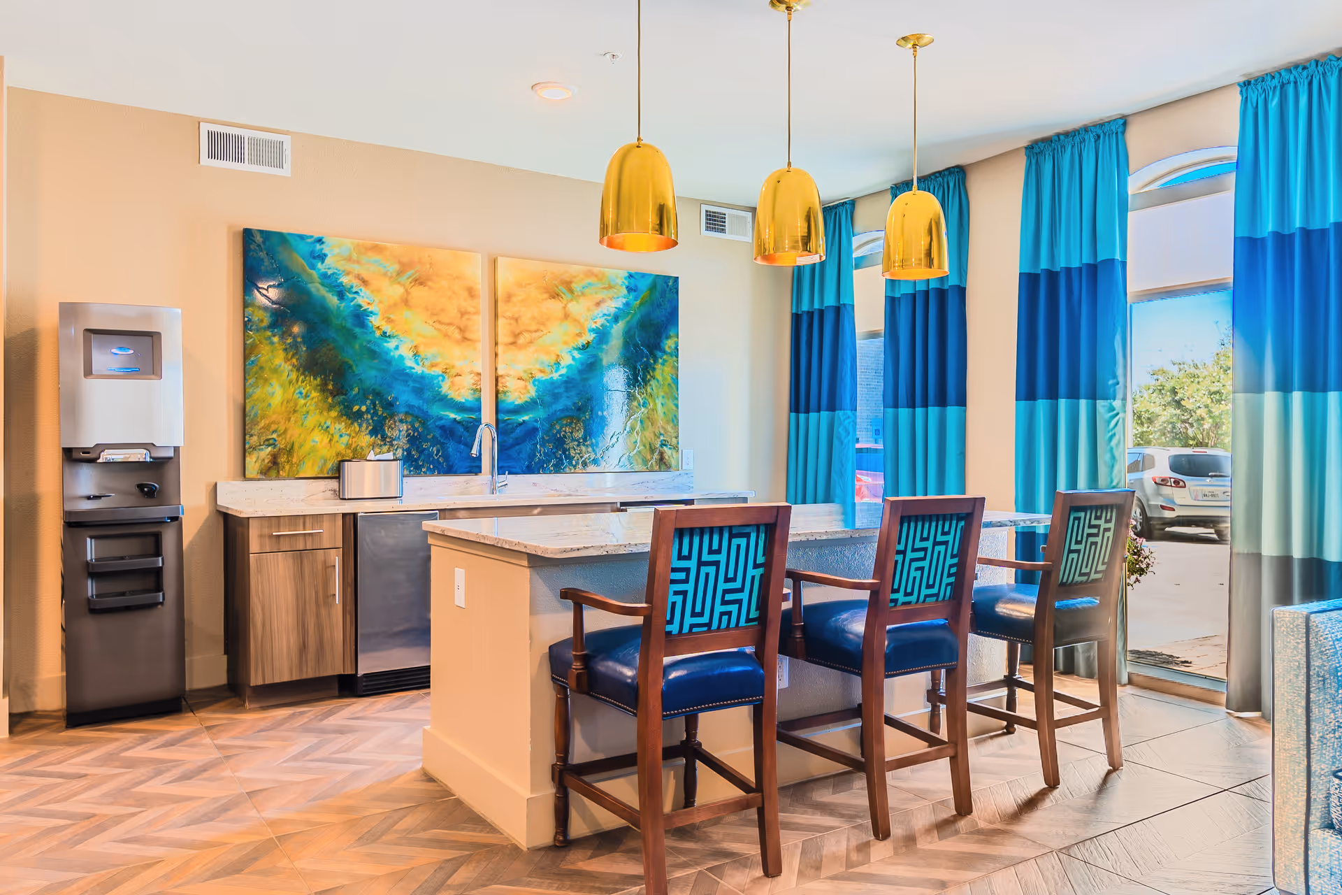 A modern kitchen area with a marble countertop island featuring three wooden chairs with blue patterned cushions. Above the island hang three gold pendant lights. Behind the island is a countertop with a sink, a tissue box, and two large abstract paintings in blue, green, and yellow tones. To the left is a stainless steel water dispenser. Large windows with blue and turquoise curtains allow natural light to fill the space, and a parking lot with cars is visible outside.