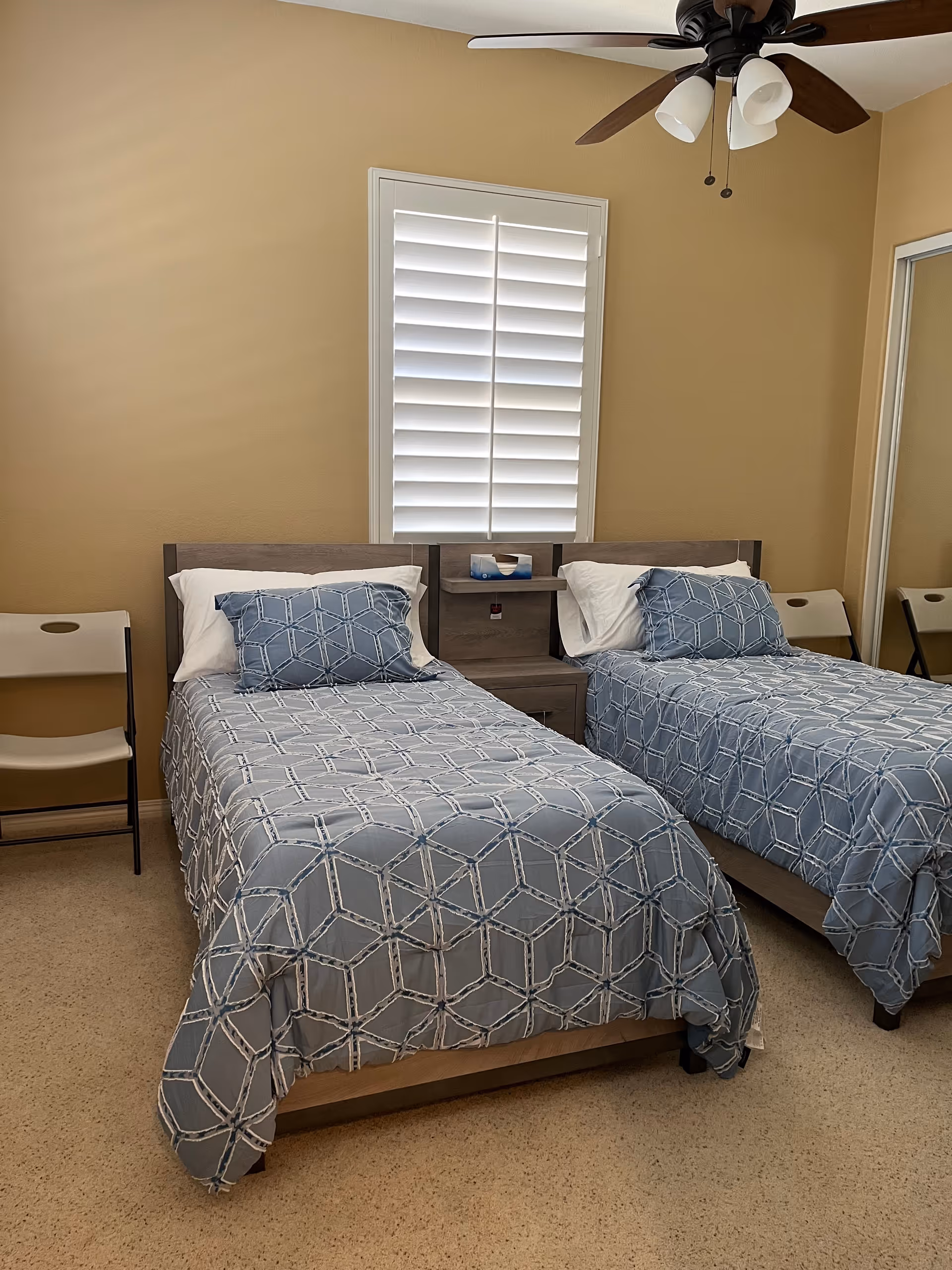A bedroom with two twin beds side by side, each with blue patterned bedspreads and matching pillows. There is a small nightstand between the beds with a tissue box on it. The room has beige walls, a window with white shutters, a ceiling fan with lights, and two beige folding chairs placed against the walls.