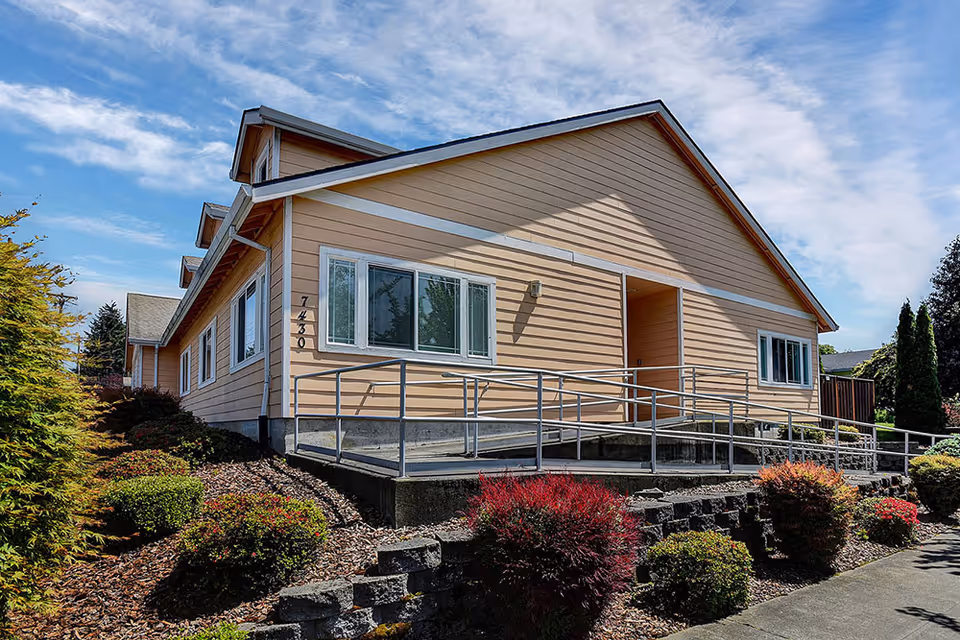Exterior view of a single-story residential building with beige siding and white trim, featuring a wheelchair accessible ramp leading to the entrance. The building is surrounded by landscaped bushes and trees under a partly cloudy blue sky.