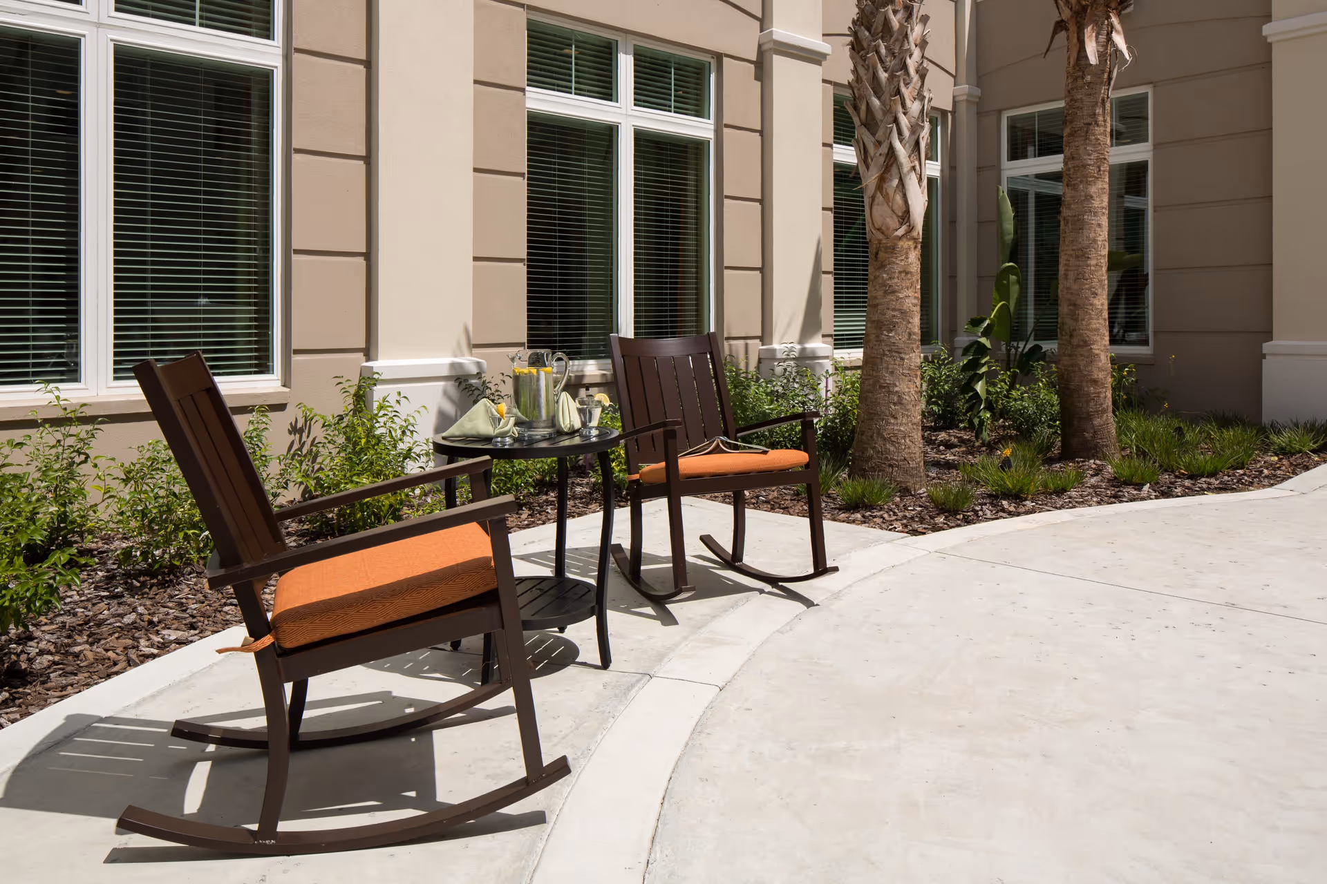 Two wooden rocking chairs with orange cushions and a small table holding drinks sit in a sunny courtyard next to a building with tall windows and palm trees.