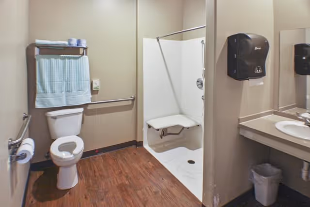 Accessible bathroom with a toilet, grab bars, a towel rack with folded towels, a walk-in shower with a fold-down seat, a sink with a countertop, a wall-mounted paper towel dispenser, and a trash bin underneath the sink. The floor has wood-like flooring and the walls are painted beige.