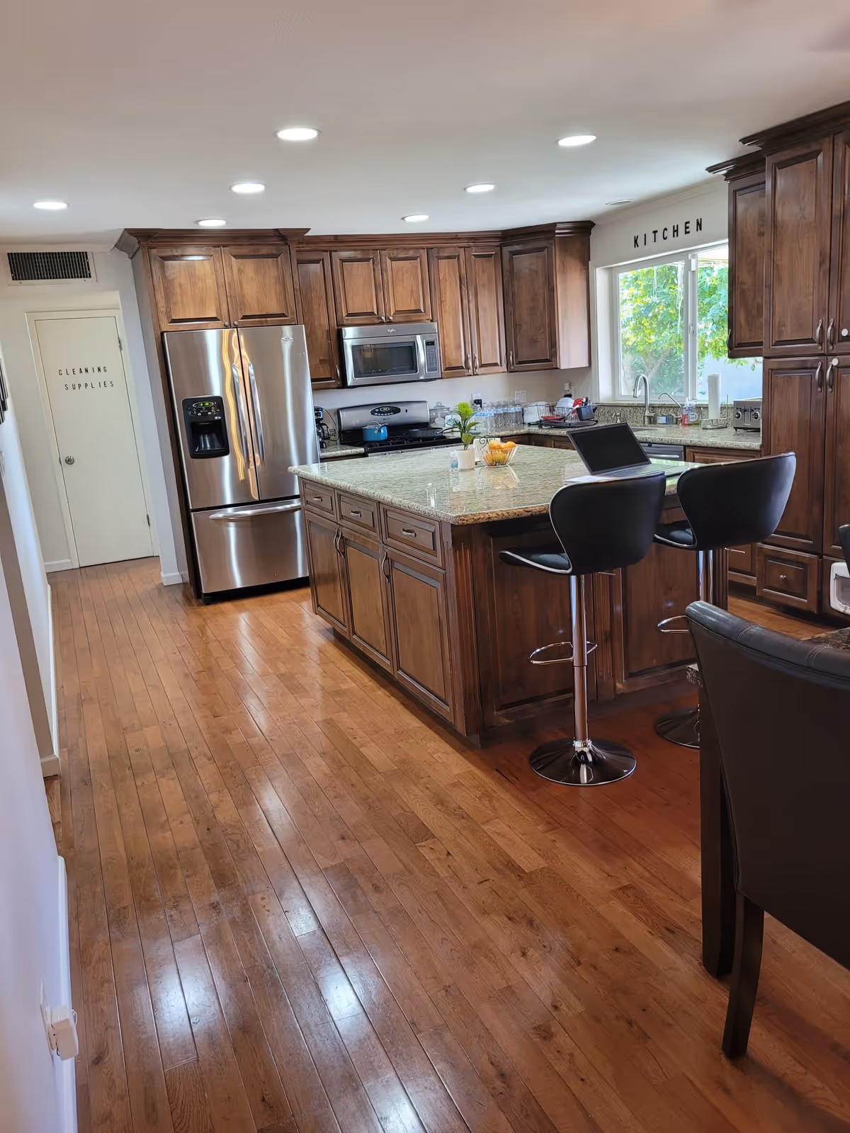 A spacious kitchen with wooden cabinets, a large granite island with two black bar stools, stainless steel refrigerator, microwave, and stove. There is a window above the sink with a view of greenery outside. The floor is wooden, and a door labeled 'Cleaning Supplies' is visible on the left side.