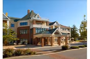 Exterior view of a multi-story senior living facility building with a covered entrance, large windows, and a mix of brick and siding. Trees and landscaping surround the building under a clear blue sky.