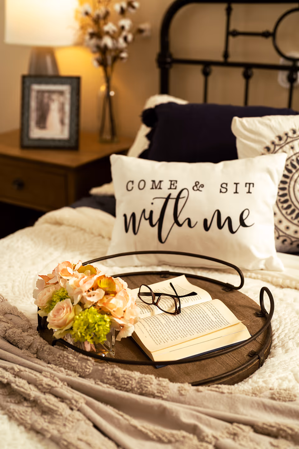 A cozy bedroom scene featuring a bed with a decorative pillow that reads 'COME & SIT with me'. On the bed is a round wooden tray holding a small bouquet of flowers, an open book, and a pair of eyeglasses. In the background, there is a nightstand with a lamp, a framed photo, and a vase with cotton stems.