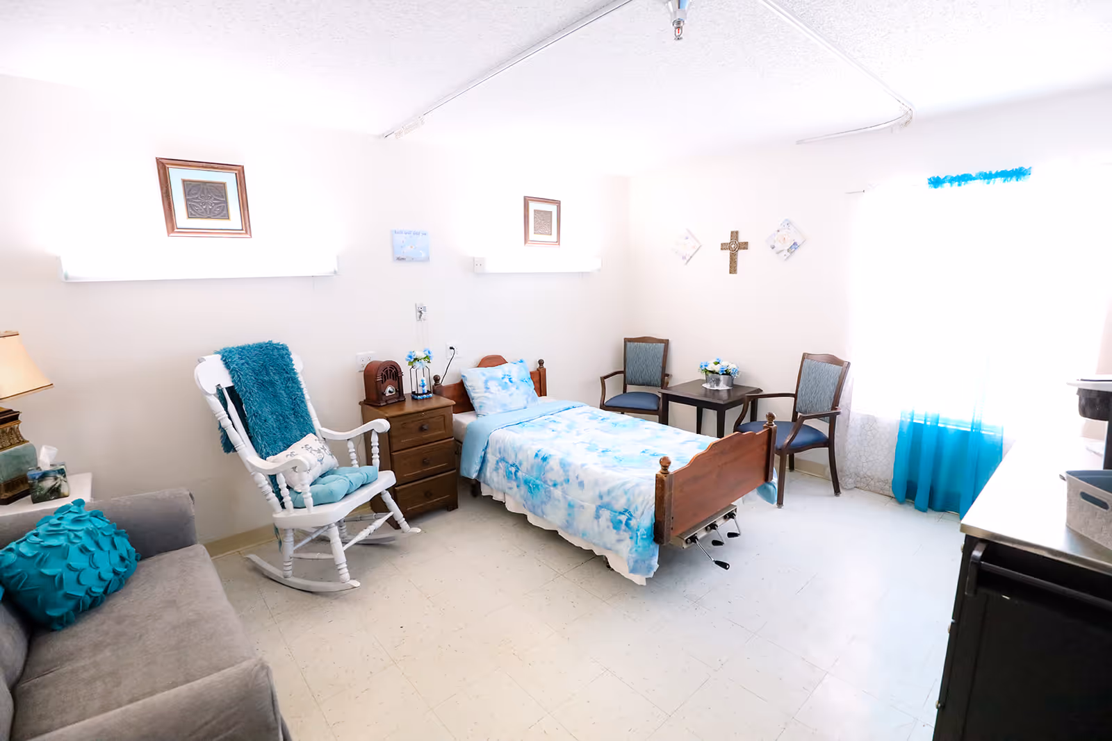 Well-lit senior bedroom with a single hospital-style bed, rocking chair, small table and seating, and teal-accented decor.