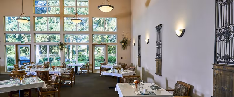 A spacious dining room with multiple tables covered in white tablecloths, set with glasses, plates, and utensils. The room features large floor-to-ceiling windows letting in natural light and offering a view of greenery outside. The walls are decorated with metal wall art and sconces, and there are hanging pendant lights from the high ceiling.