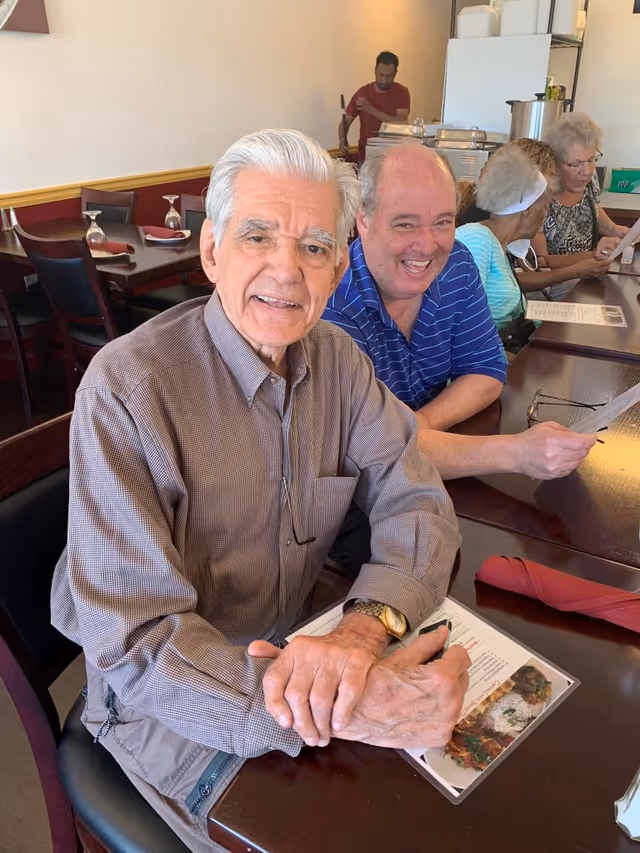 Several elderly residents sit at a dining table, smiling and looking at menus in a communal dining area.