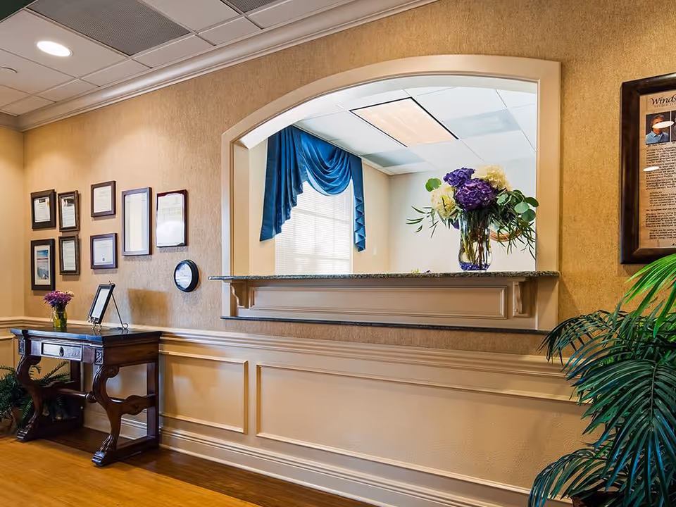 Interior view of a reception area in a senior living facility with a wooden table holding a small vase of flowers, framed certificates and documents on the wall, a large arched pass-through window with a granite ledge displaying a vase of purple and white flowers, and a potted plant in the corner.