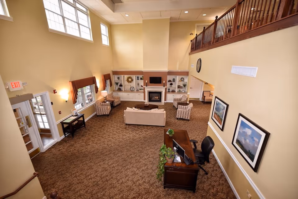 Spacious senior living facility common area with high ceilings and large windows. The room features a seating area with a sofa and four armchairs arranged around a coffee table in front of a fireplace and built-in shelves. A wooden desk with a computer and office chair is positioned near the entrance. The walls are painted light yellow, and there are framed landscape paintings on one wall. The carpet has a patterned design, and there is a balcony railing visible on the upper level.