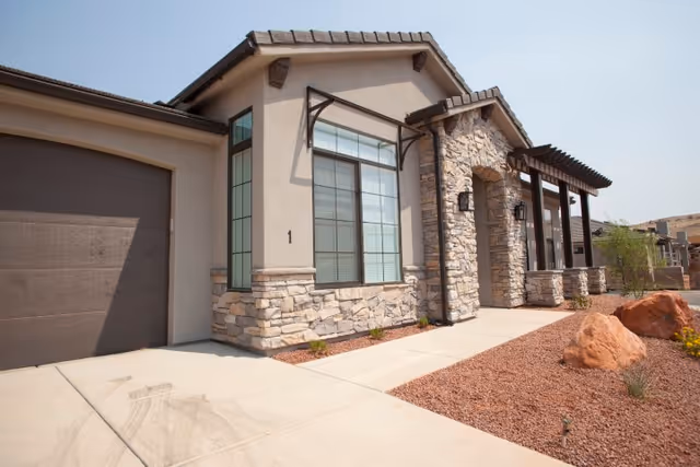 Exterior view of a single-story building with stone and stucco facade, large windows, a garage door on the left, and a covered porch supported by stone columns. The surrounding landscape features red gravel, rocks, and sparse desert plants under a clear sky.