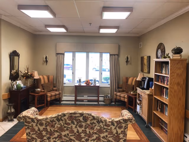 A cozy living room area with a floral patterned sofa in the foreground, two striped armchairs on either side of a window with beige curtains, a wooden bookshelf filled with books, a small table with a coffee maker, and decorative items including a clock and plants. The room has a warm and inviting atmosphere with soft lighting from ceiling fixtures.