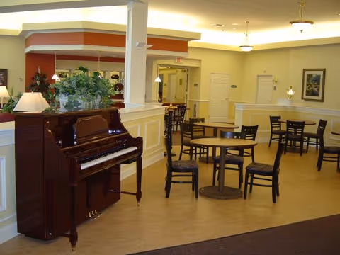 Communal dining/lounge area with round tables and chairs, a polished upright piano, and decorative plants under warm lighting.