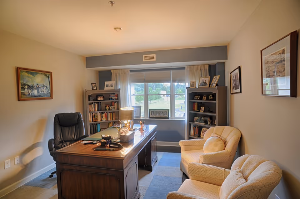 Cozy interior room with a wooden desk, office chair, two upholstered armchairs, bookshelves and a window overlooking greenery.