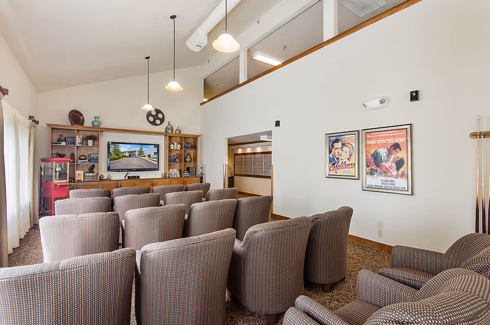 A small community theater room with rows of upholstered chairs facing a wall-mounted TV and built-in shelving, movie posters on the wall and a popcorn machine at the side.