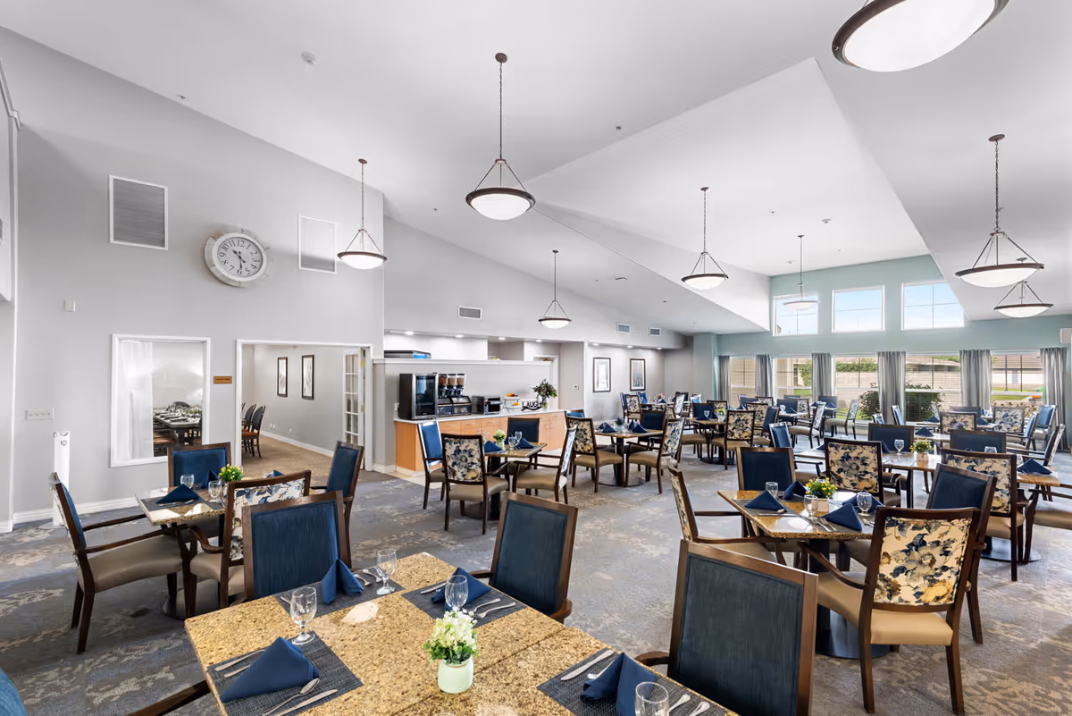 Bright and spacious dining room in a senior living facility with multiple tables set with blue napkins, glassware, and small flower centerpieces. The room features high ceilings with hanging light fixtures, large windows with curtains allowing natural light, and a coffee station area in the background.