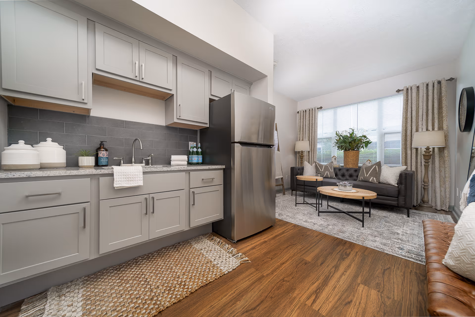 A modern open-concept living space featuring a kitchen area with gray cabinets, a stainless steel refrigerator, and a countertop with a sink and decorative items. Adjacent to the kitchen is a living room with a dark gray sofa, two round wooden coffee tables, a large window with curtains, two floor lamps, and a patterned rug on wooden flooring.