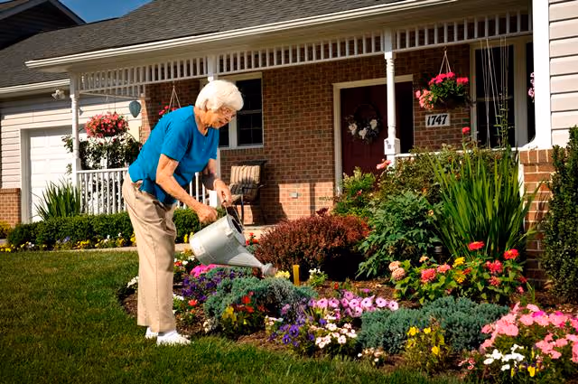 An elderly woman wearing a blue shirt and beige pants is watering a colorful flower garden in front of a brick house with a porch. The house has hanging flower baskets and the number 1747 is visible near the door.