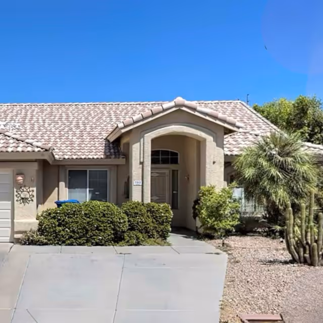 Front exterior view of a single-story house with a tiled roof, beige walls, a driveway, and desert landscaping including bushes and a cactus under a clear blue sky.