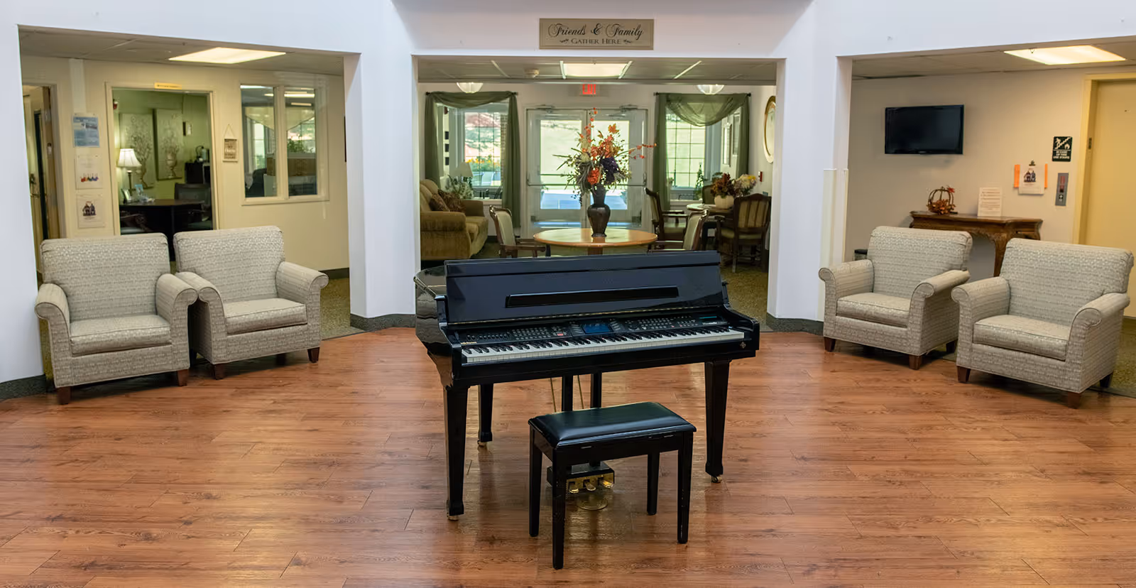 A senior living facility common area with a black piano centered on a hardwood floor and upholstered armchairs arranged along the walls.