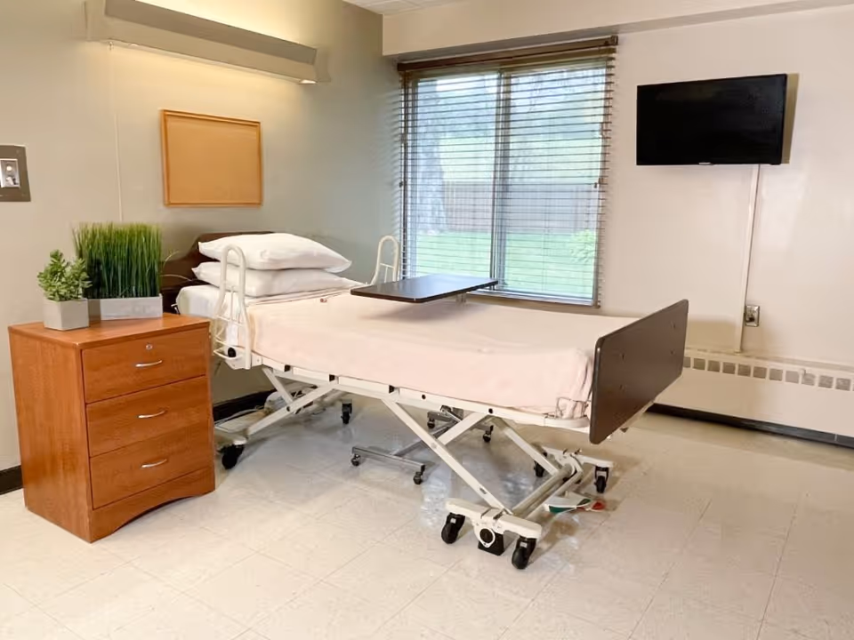 A senior living room featuring an adjustable hospital bed with overbed table, a wooden bedside dresser with plants, a wall-mounted TV, and a window with blinds.