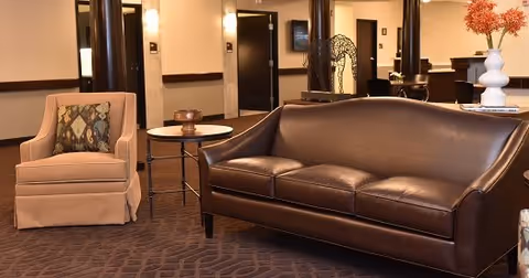 Leather sofa and upholstered armchair with side table in a carpeted lounge area of a senior living facility.