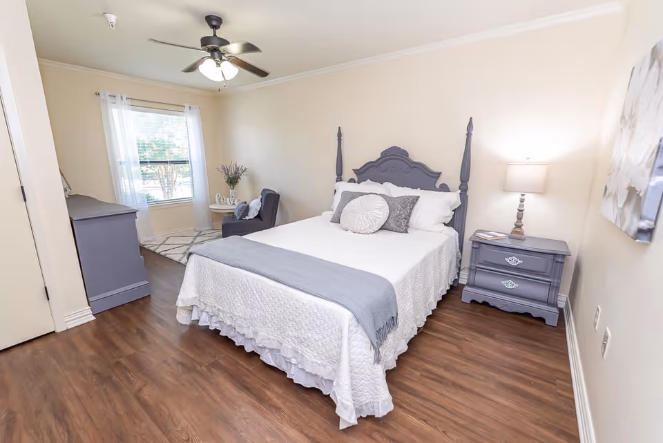 A cozy bedroom with a wooden floor, featuring a bed with a decorative headboard, white bedding, and gray pillows and throw. There is a matching gray nightstand with a lamp on the right side of the bed, a gray dresser on the left side of the room, and a small seating area with a chair and side table near a window with sheer white curtains. A ceiling fan with lights is mounted on the ceiling.