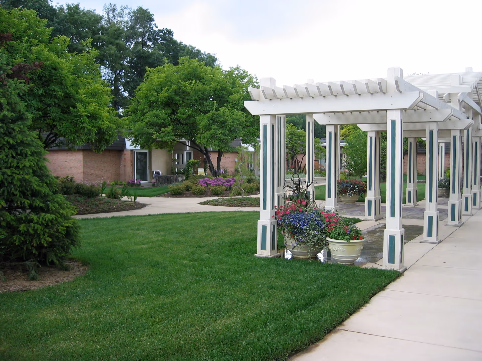 Outdoor garden area with well-maintained green grass, trees, and flower pots placed under a white pergola structure. A paved walkway runs alongside the pergola and leads to a building with brick walls and glass doors in the background.