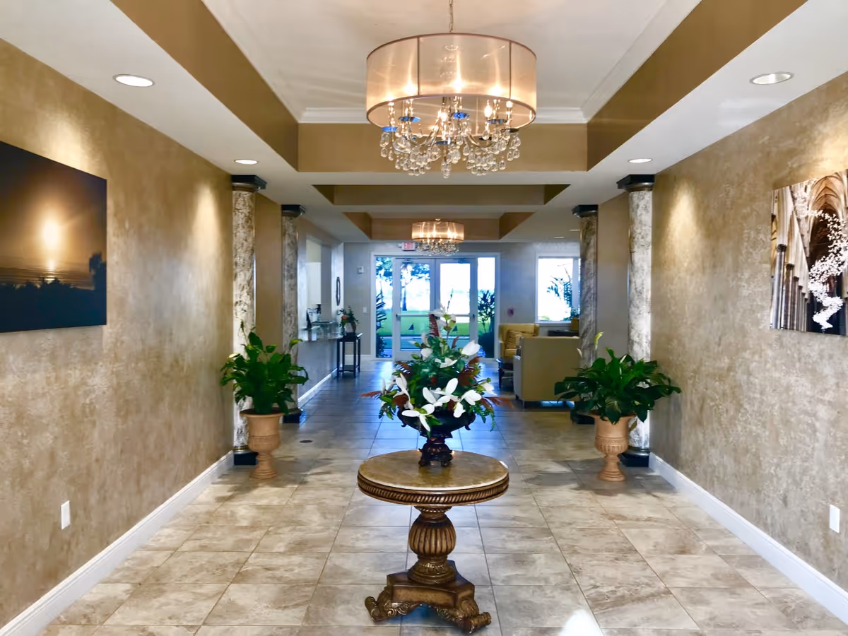 A well-lit hallway in a senior living facility with beige textured walls, tiled floor, and decorative columns. A round wooden table with a floral arrangement is centered in the hallway. Two potted plants are placed symmetrically on either side near the walls. Two chandeliers hang from the ceiling, and framed artwork is displayed on the walls. At the end of the hallway, glass doors lead to an outdoor area with visible greenery.