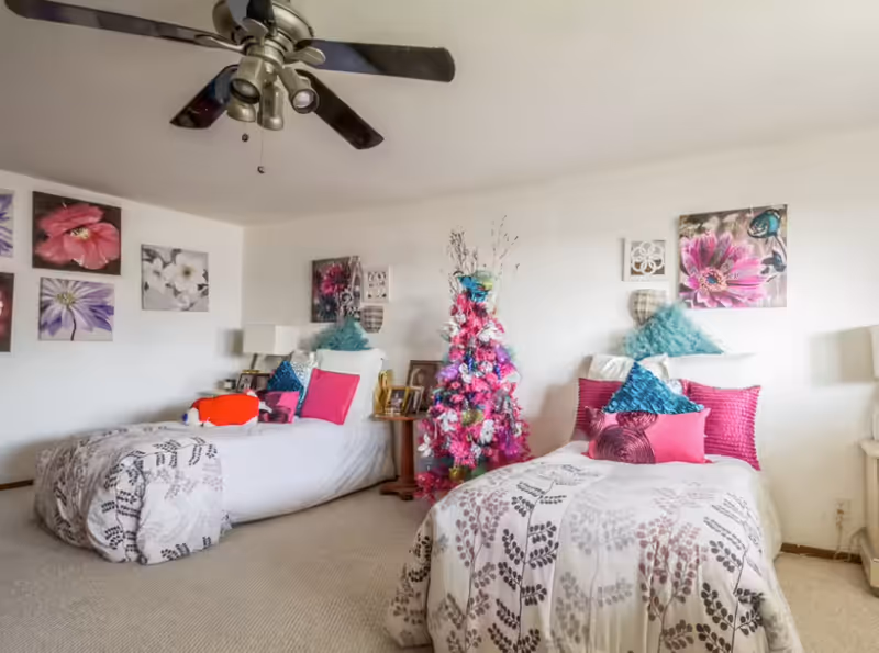 A bright bedroom with two twin beds, each adorned with white and gray patterned bedding and colorful pillows in shades of pink and teal. Between the beds is a small wooden table with framed photos and a lamp. A pink and purple decorated Christmas tree stands against the wall. The walls are decorated with floral artwork, and a ceiling fan with lights is mounted above.
