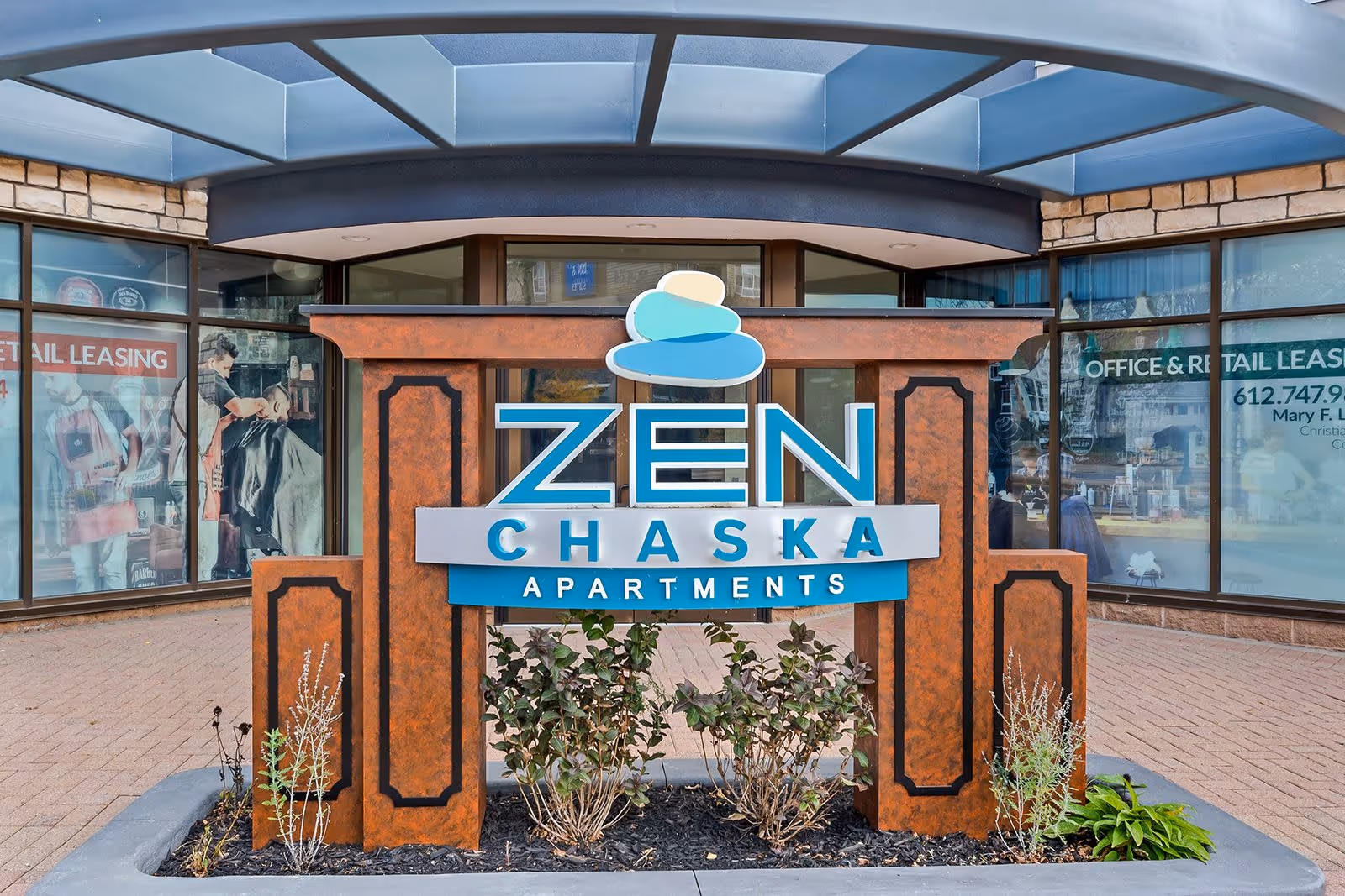 Outdoor sign for Zen Chaska Apartments with a decorative stone and blue logo, situated in front of a building with large windows displaying office and retail leasing advertisements.