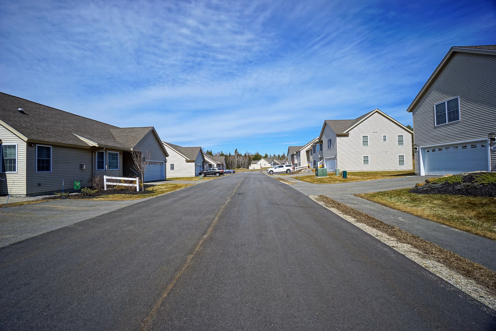 A wide paved road flanked by single-family style houses with garages on both sides under a blue sky with scattered clouds. The houses have beige siding and well-maintained lawns with some small trees and shrubs.