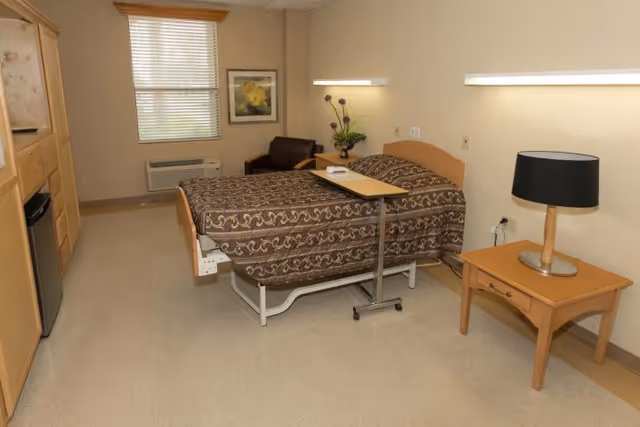 A single hospital-style bed with a patterned brown bedspread in a well-lit room. The bed has an adjustable overbed table with a phone on it. To the right of the bed is a wooden nightstand with a black lampshade. There is a window with blinds, a framed picture on the wall, a small armchair, and a wall-mounted air conditioning unit. On the left side of the room, there is built-in cabinetry with a mini refrigerator.