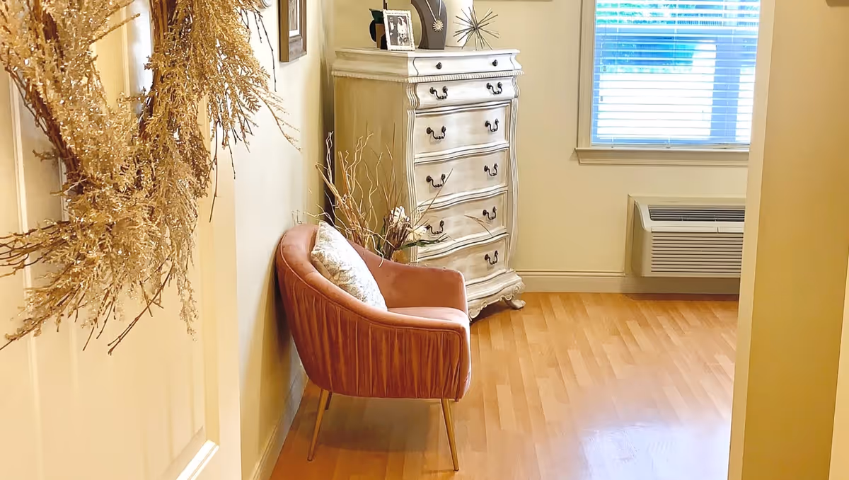 A cozy corner of a room featuring a pink upholstered chair with a decorative pillow, a vintage white chest of drawers with framed photos and decor on top, a window with blinds, and a wall-mounted air conditioning unit. A dried wreath hangs on the door to the left.
