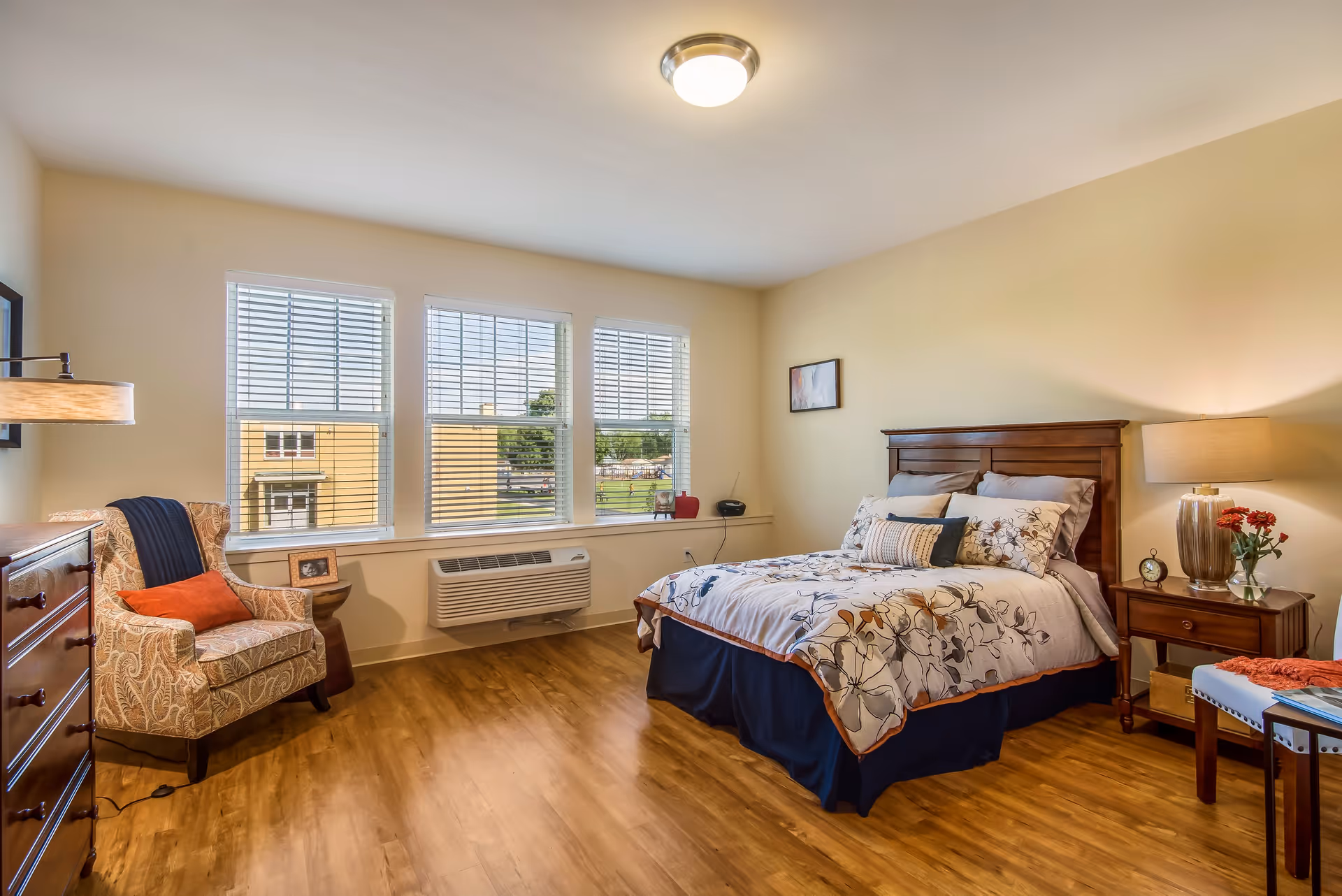 A well-lit bedroom with a large window featuring three sections with white blinds. The room has a wooden floor, a bed with floral bedding and multiple pillows, a wooden nightstand with a lamp, clock, and flowers, a patterned armchair with an orange pillow, a wooden dresser, and a small table with a chair.
