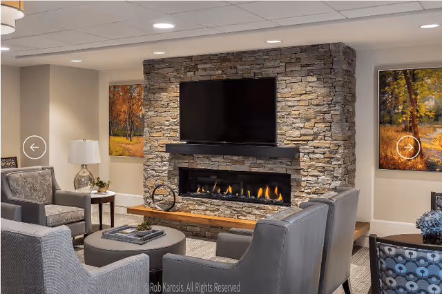 A cozy living room area featuring a stone fireplace with a modern flat-screen TV mounted above it. The fireplace is lit with a warm fire. Surrounding the fireplace are comfortable gray armchairs and a round ottoman with books and a small plant on it. The walls have framed autumn-themed paintings, and there is a side table with a lamp and decorative items.