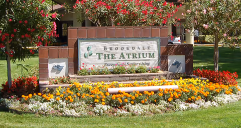 A landscaped garden area with colorful flowers including yellow, orange, red, and white blooms surrounding a brick and stone sign that reads 'Brookdale The Atrium Exceptional Senior Living'. There are trees and a building partially visible in the background.