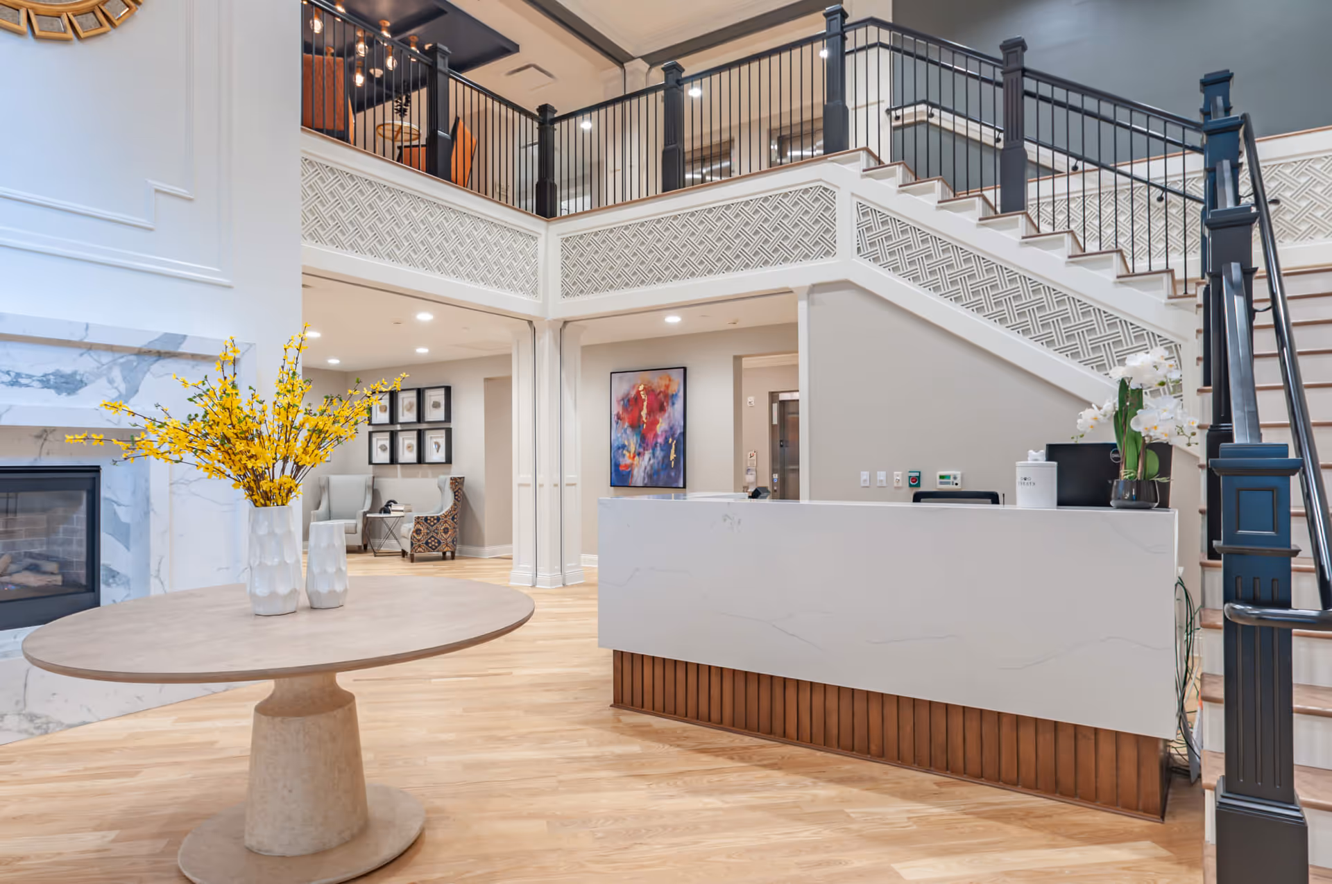 A bright and spacious reception area in Arbor Terrace Marlton featuring a modern white reception desk with a vase of white flowers, a round table with two vases holding yellow flowers, a marble fireplace, and a staircase with black railings leading to an upper level. The walls are decorated with artwork and the floor is light wood.