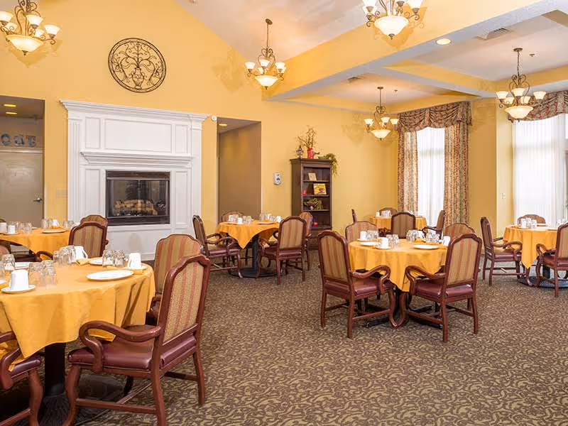 A dining room in a senior living facility with round tables covered in yellow tablecloths, each set with glasses, plates, and cups. The room features upholstered wooden chairs, a white fireplace with decorative molding, yellow walls, patterned carpet, and large windows with floral curtains. Several hanging light fixtures illuminate the space.
