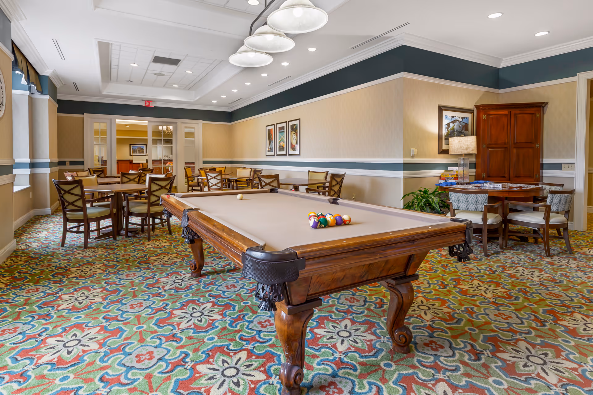 A bright, carpeted common room featuring a central pool table, multiple tables and chairs, and overhead lighting.