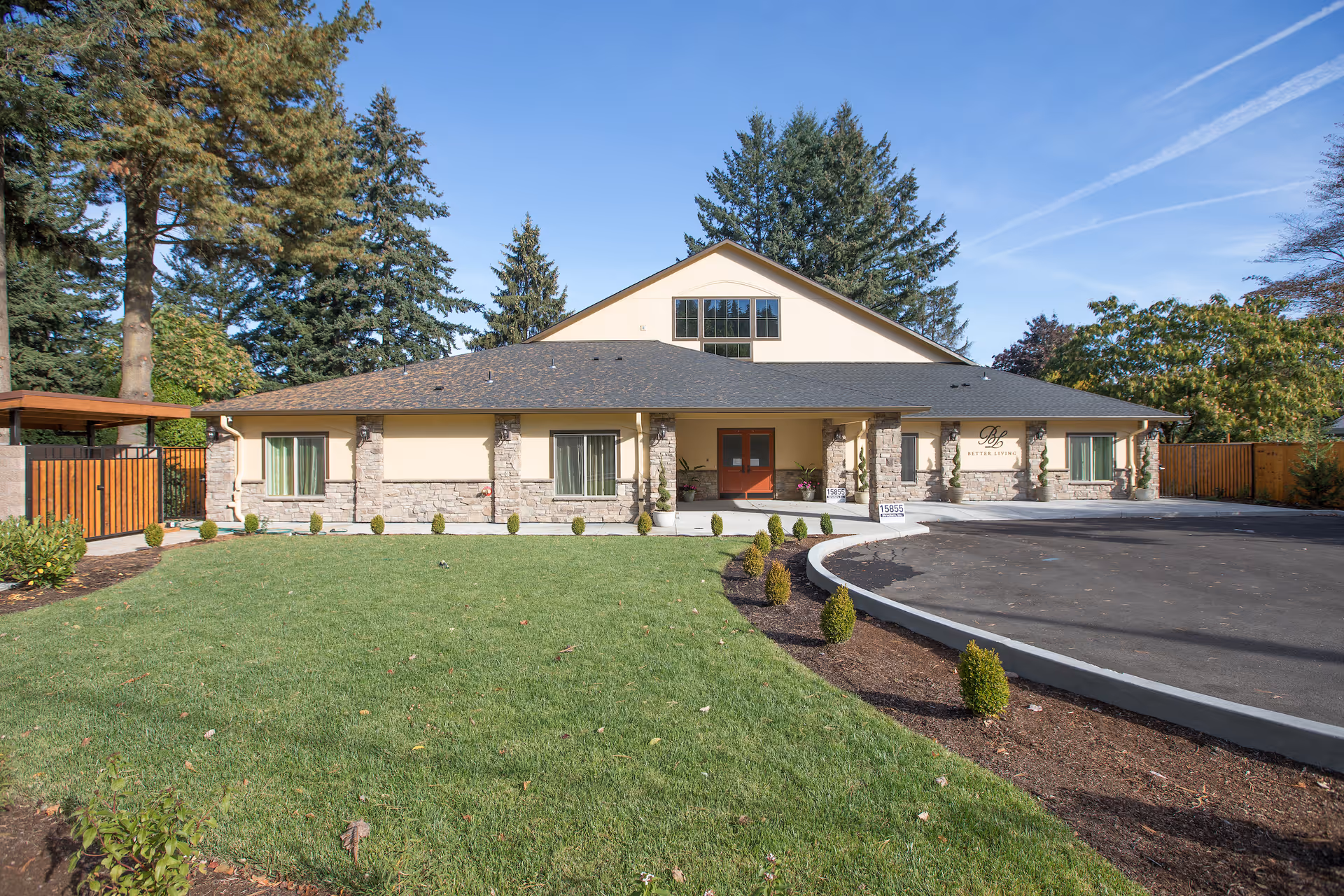 Front exterior view of a single-story building with a pitched roof, stone and beige siding, surrounded by a well-maintained lawn and small shrubs. There are large trees in the background and a paved driveway curving to the right. The building has a sign near the entrance that reads 'Better Living'.