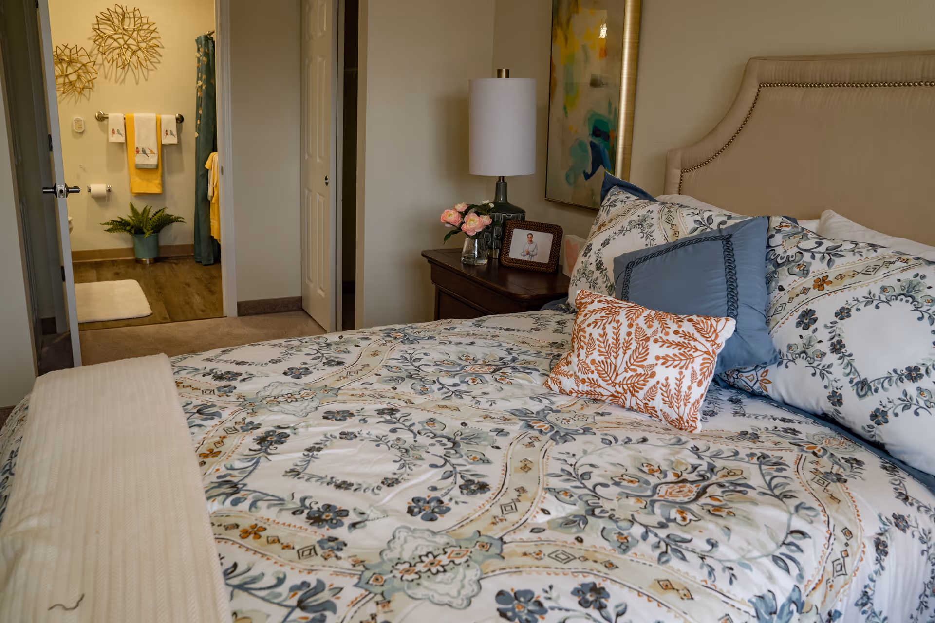 A cozy bedroom with a neatly made bed featuring patterned bedding and multiple pillows. A nightstand with a lamp, flowers, and a framed photo is beside the bed. In the background, an open door reveals a bathroom with towels hanging on the wall, a potted plant, and a shower curtain.