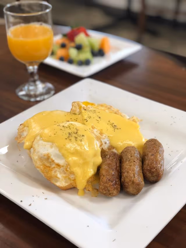 A plate of breakfast food featuring eggs topped with hollandaise sauce, three sausage links, and a glass of orange juice. In the background, there is a plate of mixed fresh fruit including melon, blueberries, and strawberries.