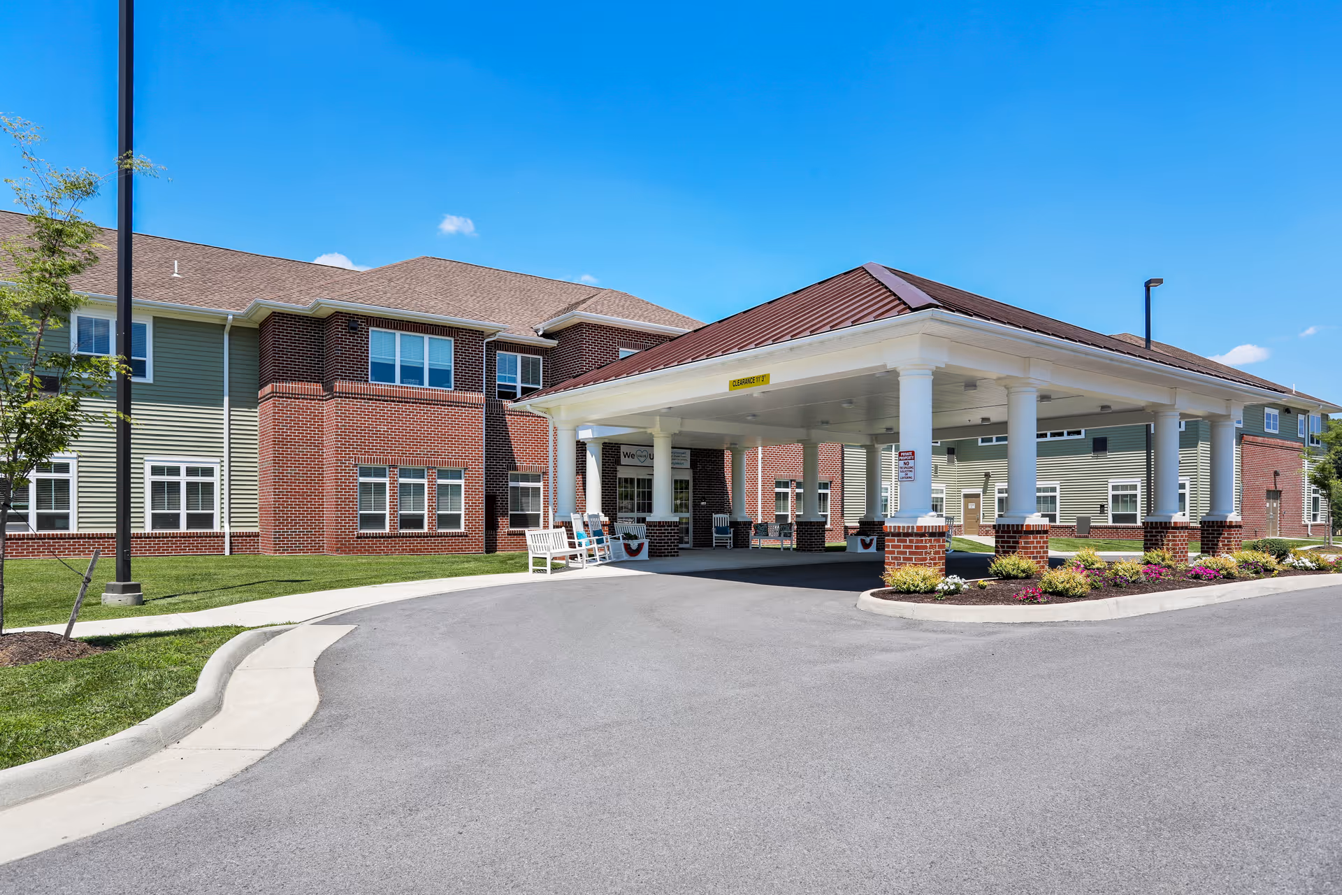 Front entrance of a senior living building with a covered porte-cochere, white columns, benches, and landscaped driveway.
