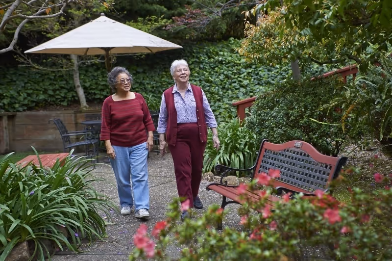 Two elderly women walking and smiling in a lush garden path surrounded by green plants and flowers, with a bench and a table with an umbrella in the background.